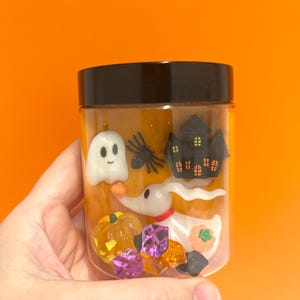 May include: A clear jar with a black lid filled with Halloween decorations. The jar contains a white ghost, a black spider, a haunted house, a dog-like character, pumpkins, and colourful gems. The jar is held against an orange background.