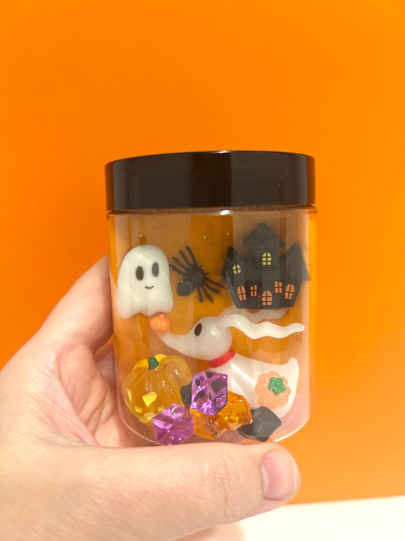 May include: A clear jar with a black lid filled with Halloween decorations. The jar contains a white ghost, a black spider, a haunted house, a dog-like character, pumpkins, and colourful gems. The jar is held against an orange background.