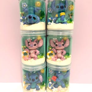 Puede incluir: Seis frascos de pl&aacute;stico transparente llenos de dioramas con tem&aacute;tica de playa. Cada frasco presenta una figura diferente de Stitch o Angel, rodeada de palmeras en miniatura, flores y un salvavidas. El fondo es de color verde azulado, y los frascos est&aacute;n apilados.