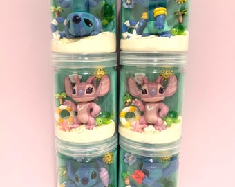 May include: Six clear cylindrical jars containing miniature dioramas featuring blue and pink alien characters. Each jar includes a sandy base, a small green turtle, a life preserver, a palm tree, a yellow sun, and a blue flower, creating a beach scene.