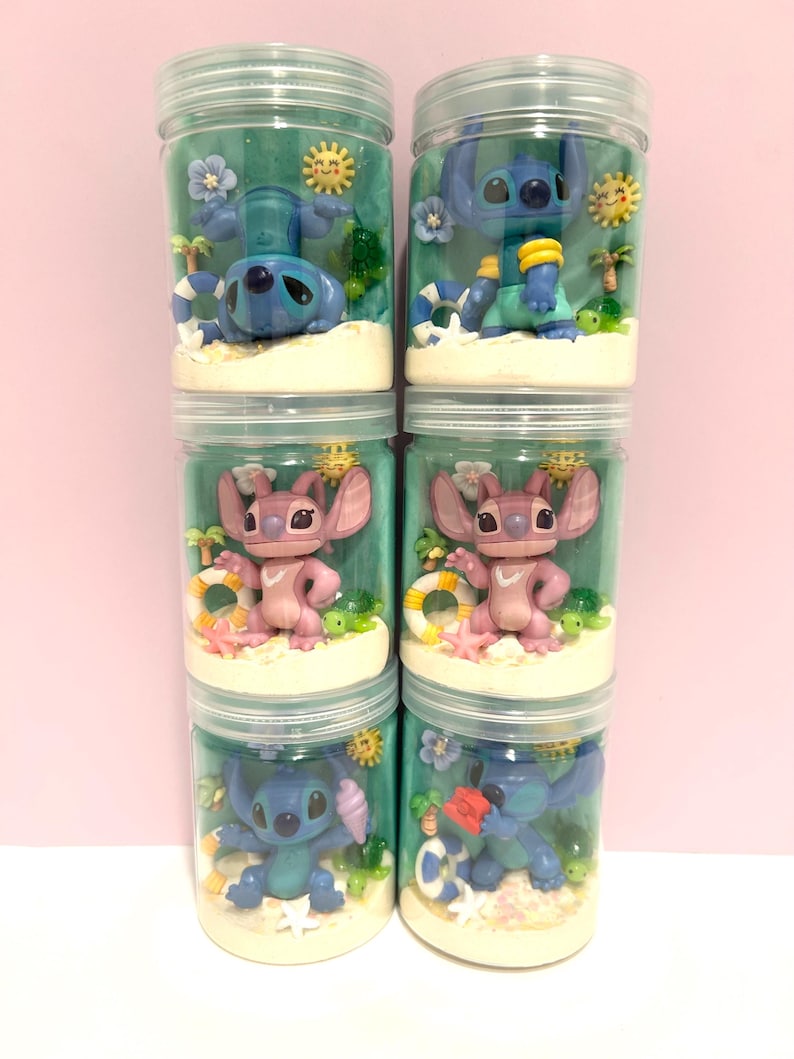 Puede incluir: Seis frascos de pl&aacute;stico transparente llenos de dioramas con tem&aacute;tica de playa. Cada frasco presenta una figura diferente de Stitch o Angel, rodeada de palmeras en miniatura, flores y un salvavidas. El fondo es de color verde azulado, y los frascos est&aacute;n apilados.