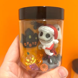 May include: A clear jar with a black lid filled with Halloween-themed figurines. The jar contains a Jack Skellington figure in a Santa Claus outfit, a ghost, a pumpkin, and a haunted house. Assorted sweets and gems are also inside.
