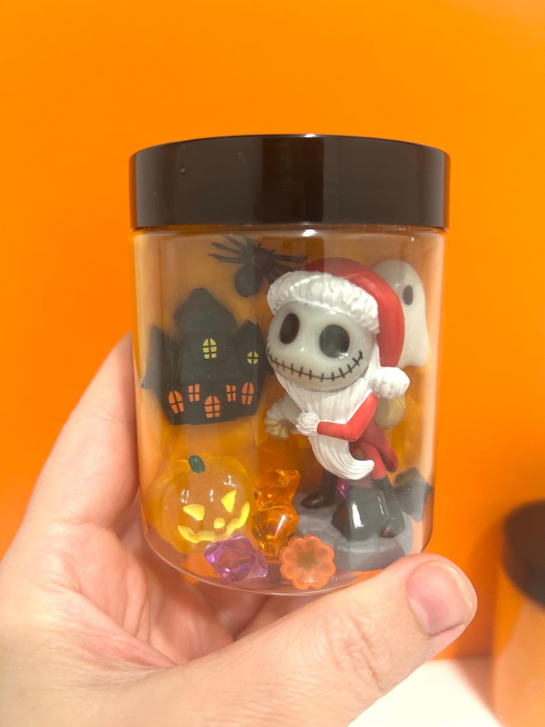 May include: A clear jar with a black lid filled with Halloween-themed figurines. The jar contains a Jack Skellington figure in a Santa Claus outfit, a ghost, a pumpkin, and a haunted house. Assorted sweets and gems are also inside.