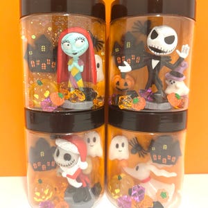 May include: Four clear plastic jars with black lids, each filled with Halloween-themed figurines and glitter. The jars feature characters like Sally, Jack Skellington, ghosts, pumpkins, and miniature haunted houses. The background is orange.