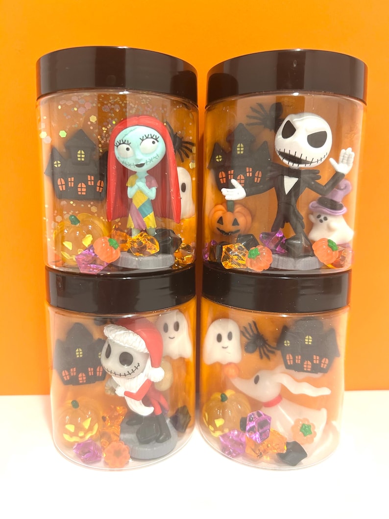 May include: Four clear plastic jars with black lids, each filled with Halloween-themed figurines and glitter. The jars feature characters like Sally, Jack Skellington, ghosts, pumpkins, and miniature haunted houses. The background is orange.