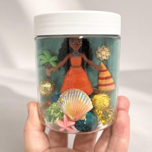 May include: A clear plastic jar with a white lid containing a small doll with long dark hair wearing an orange outfit, surrounded by miniature items including a palm tree, a sailboat, a seashell, a pink starfish, and various colorful gems.
