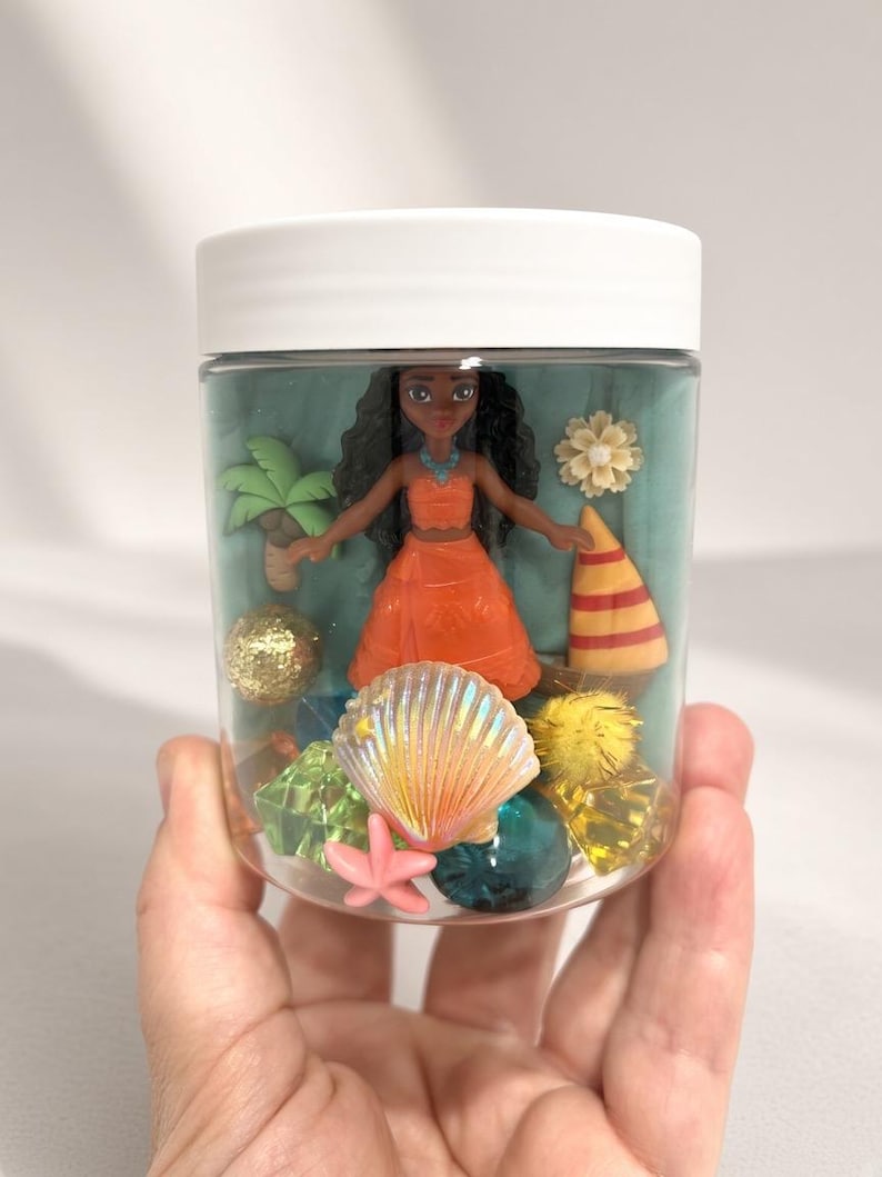 May include: A clear plastic jar with a white lid containing a small doll with long dark hair wearing an orange outfit, surrounded by miniature items including a palm tree, a sailboat, a seashell, a pink starfish, and various colorful gems.