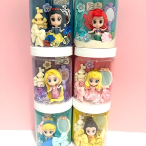 Peut inclure: Six bocaux en plastique transparent avec des couvercles blancs, chacun contenant une figurine de princesse miniature. Les bocaux présentent Blanche-Neige, Ariel, Raiponce, Aurore, Cendrillon et une sixième princesse. Chaque bocal a un fond unique et des éléments décoratifs.