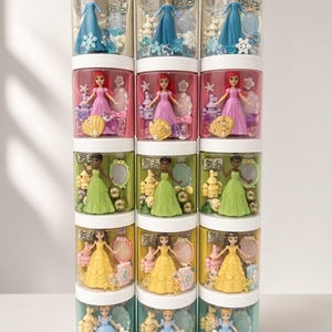 May include: A vertical stack of fifteen clear cylindrical containers with white lids, each holding a small princess figurine, a castle, a mirror, and themed decorative beads. The sets feature characters in blue, pink, green, yellow, and light blue dresses.