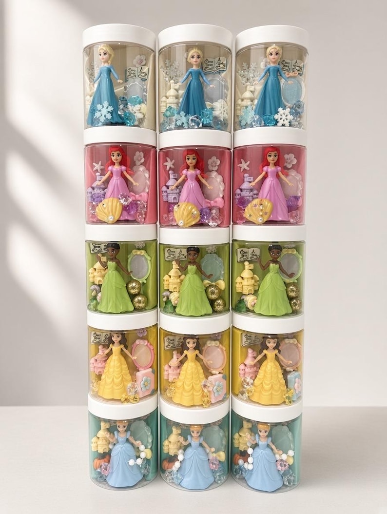 May include: A vertical stack of fifteen clear cylindrical containers with white lids, each holding a small princess figurine, a castle, a mirror, and themed decorative beads. The sets feature characters in blue, pink, green, yellow, and light blue dresses.