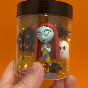 May include: A clear plastic jar with a black lid, filled with glitter and Halloween-themed figurines. The jar contains a Sally figure, a ghost, a pumpkin, a spider, and a haunted house. The background is a solid orange.