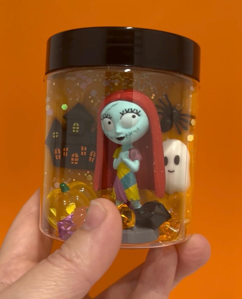 May include: A clear plastic jar with a black lid, filled with glitter and Halloween-themed figurines. The jar contains a Sally figure, a ghost, a pumpkin, a spider, and a haunted house. The background is a solid orange.