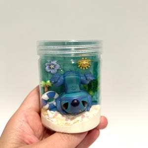 Puede incluir: Un tarro de pl&aacute;stico transparente que contiene una figura azul de Stitch en una escena de playa. La escena incluye un salvavidas azul y blanco, un sol amarillo y una flor azul. El tarro est&aacute; lleno de un l&iacute;quido azul y arena en el fondo.
