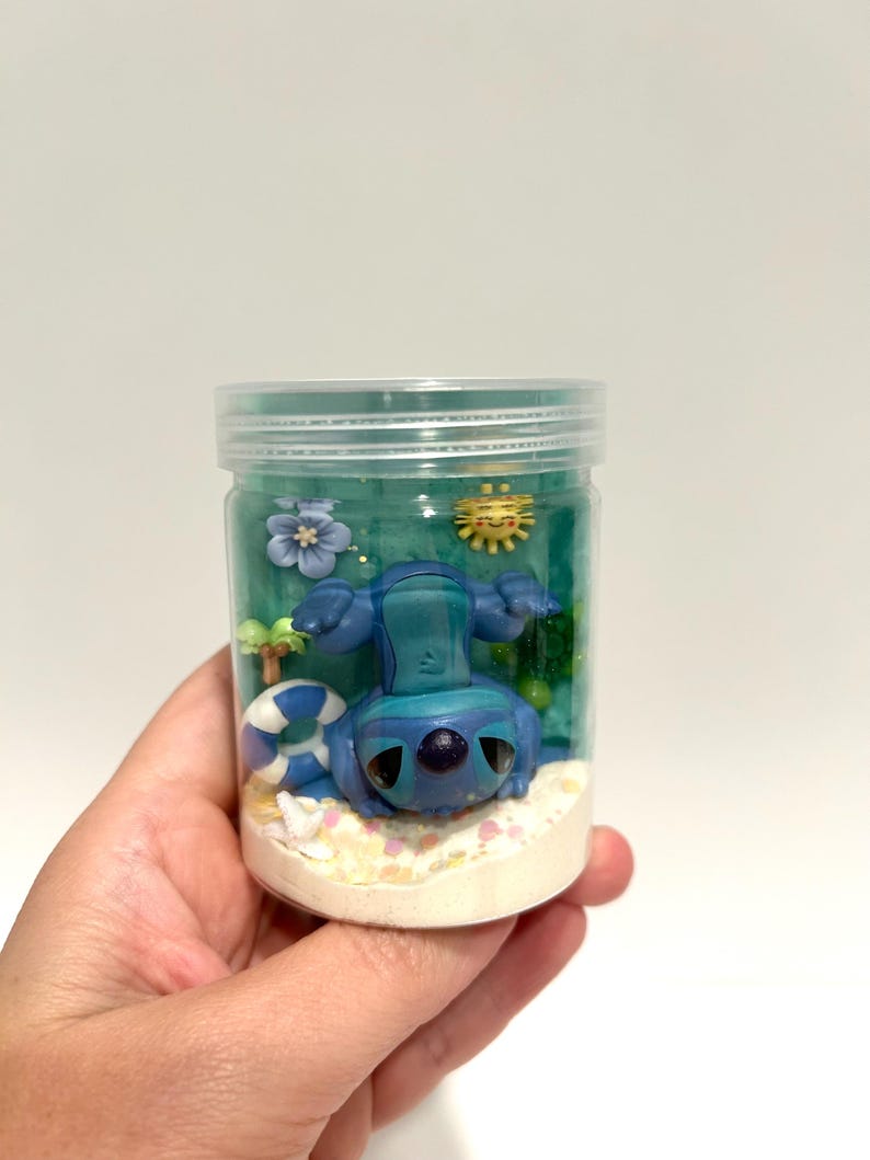 Puede incluir: Un tarro de pl&aacute;stico transparente que contiene una figura azul de Stitch en una escena de playa. La escena incluye un salvavidas azul y blanco, un sol amarillo y una flor azul. El tarro est&aacute; lleno de un l&iacute;quido azul y arena en el fondo.