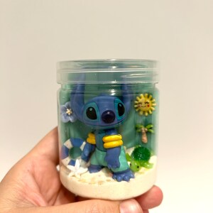 Puede incluir: Un frasco transparente lleno de una escena de playa con un personaje alien&iacute;gena azul con detalles amarillos, una peque&ntilde;a tortuga verde, una palmera y un sol amarillo. El frasco tiene una tapa transparente y est&aacute; lleno de arena y gel azul.