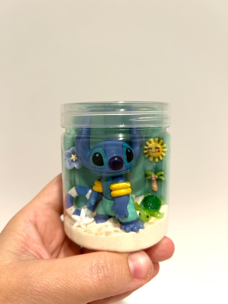 Puede incluir: Un frasco transparente lleno de una escena de playa con un personaje alien&iacute;gena azul con detalles amarillos, una peque&ntilde;a tortuga verde, una palmera y un sol amarillo. El frasco tiene una tapa transparente y est&aacute; lleno de arena y gel azul.