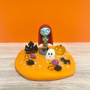 May include: A Halloween diorama with a Sally figurine, red hair and patchwork dress. The scene includes a black haunted house, a carved pumpkin, a white ghost, a black spider, and colourful gems on an orange base.