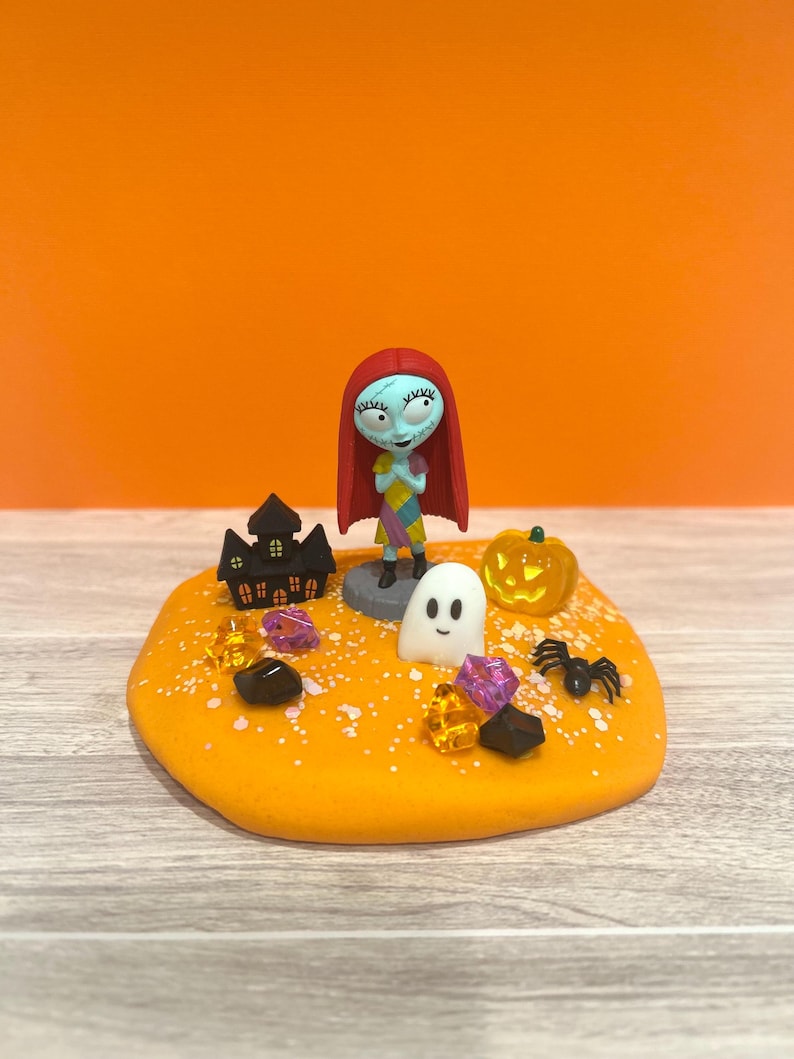 May include: A Halloween diorama with a Sally figurine, red hair and patchwork dress. The scene includes a black haunted house, a carved pumpkin, a white ghost, a black spider, and colourful gems on an orange base.