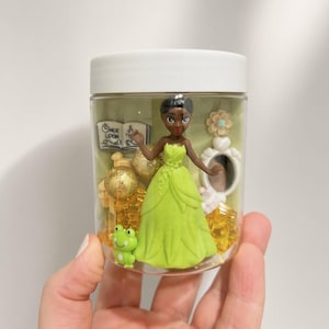 May include: A clear plastic jar with a white lid containing a figurine of a person in a lime green dress, a small green frog, gold glitter spheres, a white mirror, a flower, and a book with the text Once Upon A Time printed on the cover.
