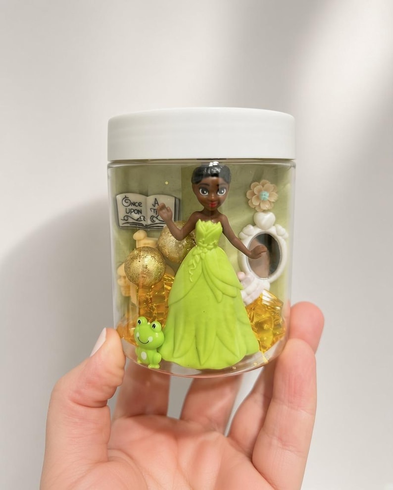 May include: A clear plastic jar with a white lid containing a figurine of a person in a lime green dress, a small green frog, gold glitter spheres, a white mirror, a flower, and a book with the text Once Upon A Time printed on the cover.
