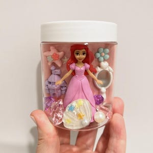 May include: A clear plastic jar with a white lid containing a small red-haired doll in a pink dress, a purple castle figurine, a white hand mirror, a pink starfish, a white seashell with colorful stars, and various translucent purple and clear gems.