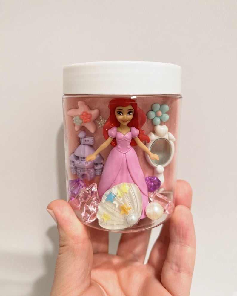 May include: A clear plastic jar with a white lid containing a small red-haired doll in a pink dress, a purple castle figurine, a white hand mirror, a pink starfish, a white seashell with colorful stars, and various translucent purple and clear gems.