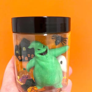 May include: A clear plastic jar with a black lid filled with Halloween decorations. Inside are a green Oogie Boogie figure, a black spider, a white ghost, a pumpkin, and a haunted house silhouette. The background is a solid orange.