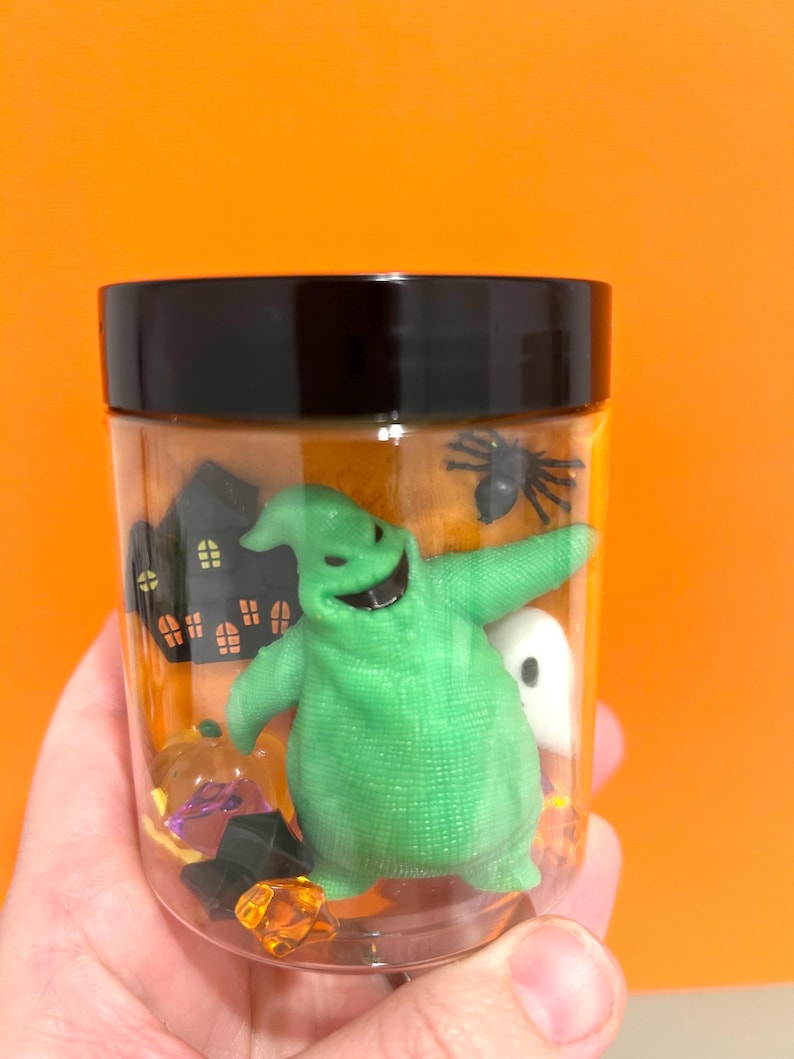 May include: A clear plastic jar with a black lid filled with Halloween decorations. Inside are a green Oogie Boogie figure, a black spider, a white ghost, a pumpkin, and a haunted house silhouette. The background is a solid orange.