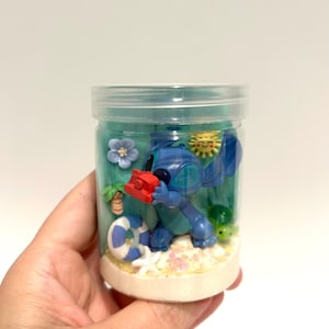 Puede incluir: Un tarro de pl&aacute;stico transparente que contiene una escena de playa con una figura azul de Stitch sosteniendo un objeto rojo. La escena incluye un salvavidas azul y blanco, una estrella de mar y una peque&ntilde;a tortuga. El tarro est&aacute; lleno de l&iacute;quido azul y arena.
