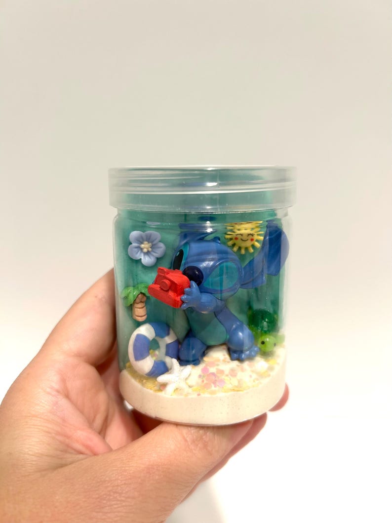 Puede incluir: Un tarro de pl&aacute;stico transparente que contiene una escena de playa con una figura azul de Stitch sosteniendo un objeto rojo. La escena incluye un salvavidas azul y blanco, una estrella de mar y una peque&ntilde;a tortuga. El tarro est&aacute; lleno de l&iacute;quido azul y arena.