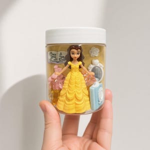 May include: A clear plastic jar with a white lid containing a small Belle figurine in a yellow dress, a miniature book with the text Once Upon A Time, a white flower, a white mirror, a light blue book, and several translucent pink and yellow gems.