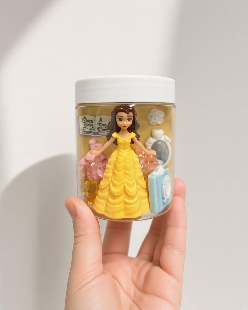 May include: A clear plastic jar with a white lid containing a small Belle figurine in a yellow dress, a miniature book with the text Once Upon A Time, a white flower, a white mirror, a light blue book, and several translucent pink and yellow gems.