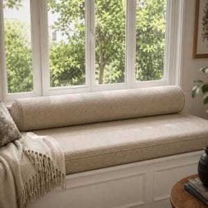 May include: A long, cylindrical bolster pillow in a textured beige fabric sits atop a matching rectangular bench cushion. A beige throw blanket with fringe hangs over the side of the white window seat, positioned in front of a large window.