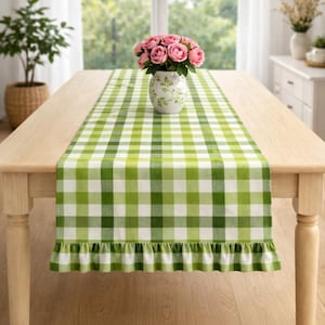May include: A light wooden table with a green and white checkered table runner. A vase with pink roses sits in the center of the table. The runner has a ruffled edge. A potted plant is in the background.