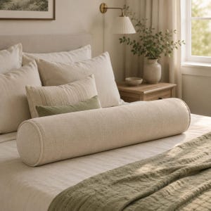 May include: A long, cylindrical bolster pillow in a neutral beige linen-like fabric with piped edges. The pillow is positioned horizontally across the front of a bed layered with various rectangular pillows and a textured olive green throw blanket.