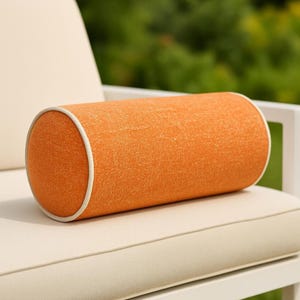 May include: An orange cylindrical bolster pillow with a white trim, resting on a cream-colored cushion. The pillow has a textured surface and is designed for comfort and support. The outdoor setting suggests patio or garden use.