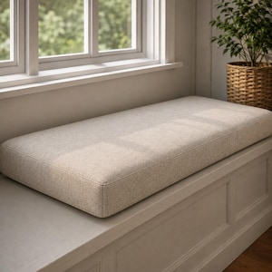 May include: A rectangular, beige window seat cushion with a textured fabric cover. The cushion is placed on a white window seat, near a window and a potted plant in a woven basket.
