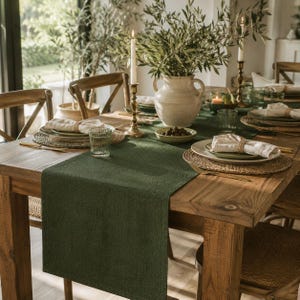 Può includere: Un runner da tavola rettangolare in tessuto verde scuro posizionato su un tavolo da pranzo in legno. Il runner presenta una trama testurizzata ed è collocato al centro, fungendo da base per le stoviglie con tovagliette intrecciate, tovaglioli e candele.