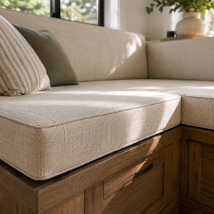 May include: A corner bench featuring a natural wood base with integrated storage drawers and thick, light beige textured fabric cushions. The seating area includes a striped throw pillow and a solid olive green pillow, providing a neutral aesthetic.