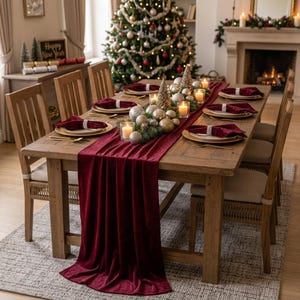 Op de afbeelding: Een houten eettafel gedekt voor een feestelijke maaltijd met een dieprode fluwelen tafelloper, bijpassende rode fluwelen servetten met kralenringen, witte borden, gouden bestek en een middelpunt van zilveren en gouden kerstballen.