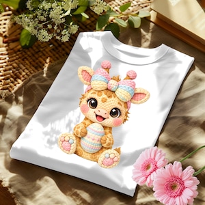 May include: White t-shirt featuring a cartoon giraffe holding a decorated Easter egg. The giraffe has a pink, blue, and yellow bow and is sitting on the shirt. Pink flowers are in the bottom right corner.