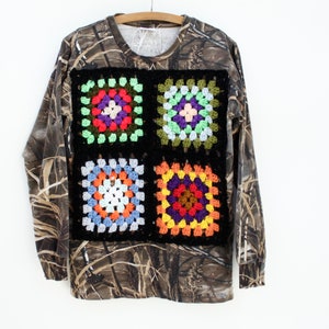 May include: A long-sleeved shirt with a camouflage pattern and four colorful crocheted squares on the front.
