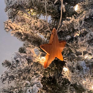 May include: A wooden star Christmas ornament with a natural wood grain finish hangs from a string against a frosted evergreen tree. The star is illuminated by the warm glow of Christmas lights, creating a festive holiday scene.