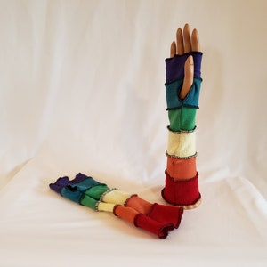 May include: A pair of rainbow fingerless gloves with a contrasting red trim. The gloves are made of a soft, knitted fabric and feature a variety of colors, including red, orange, yellow, green, blue, and purple.