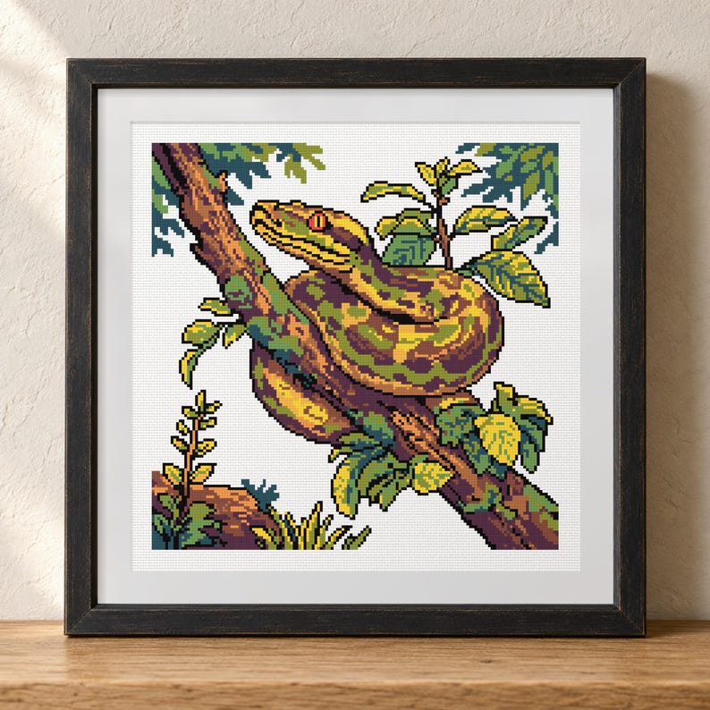 May include: A framed cross-stitch embroidery featuring a pixel art style snake coiled around a tree branch. The snake has green and yellow scales with a red eye. The scene includes green leaves and a brown branch, all set within a dark wooden frame.