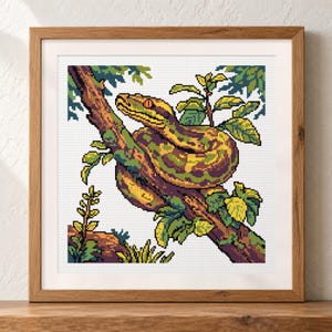 May include: A square cross-stitch pattern featuring a pixel art style snake coiled around a tree branch. The snake has green and yellow scales with a red eye. The scene includes green leaves and a brown branch, all framed in a light wood frame.