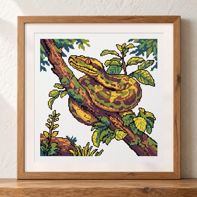 May include: A square cross-stitch pattern featuring a pixel art style snake coiled around a tree branch. The snake has green and yellow scales with a red eye. The scene includes green leaves and a brown branch, all framed in a light wood frame.