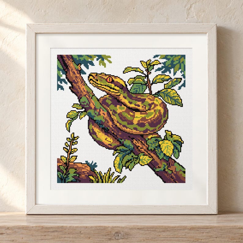 May include: A square framed cross-stitch embroidery featuring a pixel art style snake coiled around a tree branch. The snake has yellow, green, and brown scales with a bright orange eye. Green leaves and foliage surround the branch against a white background.
