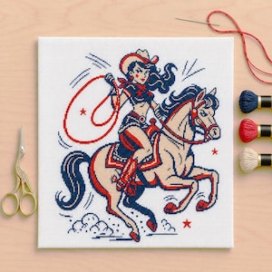 Pode incluir: Uma bordado em ponto cruz de uma cowgirl montada a cavalo, em vermelho, azul marinho e bege. A cowgirl usa um chapéu e segura um laço. O cavalo galopa, com estrelas e nuvens.