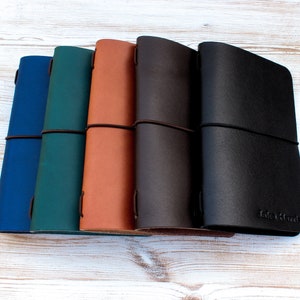 May include: A collection of leather journals in various colors: blue, teal, tan, dark brown, and black. Each journal is closed with an elastic band. The black journal has the text "Lake Harada" embossed on the front.